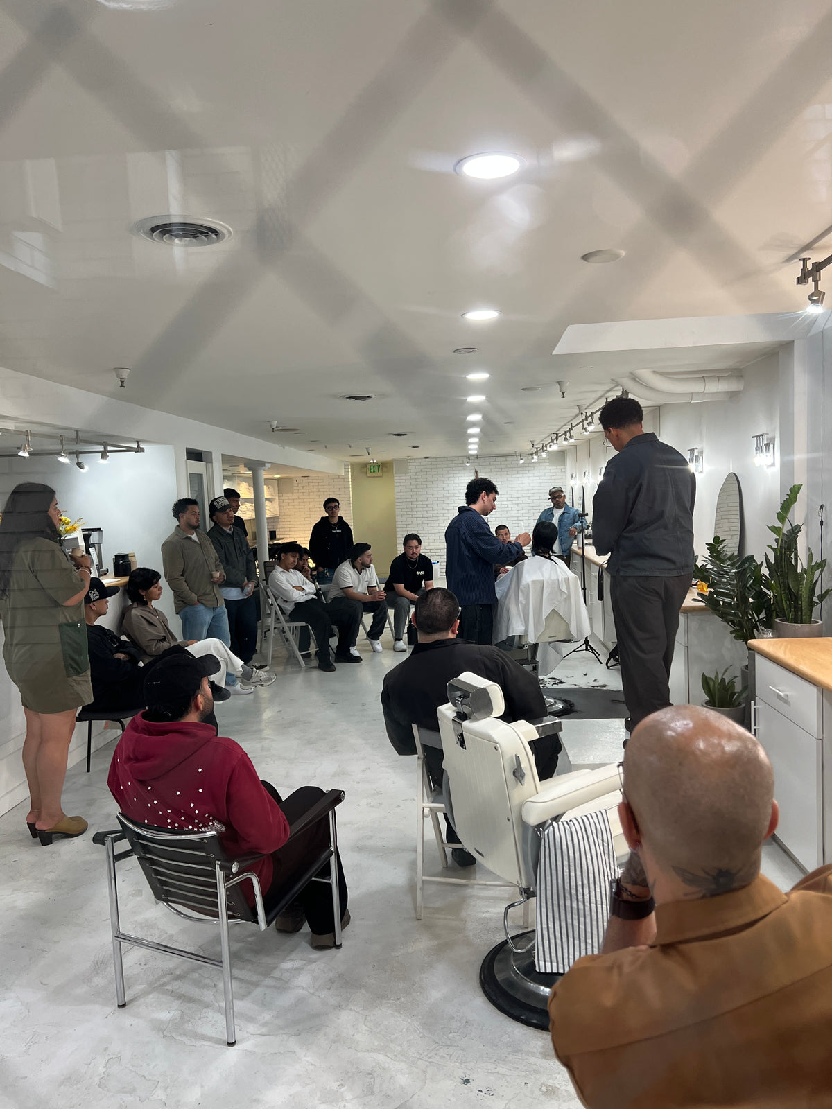 A barbershop interior with multiple people gathered around barber chairs, watching a demonstration. The room has white walls and ceiling, bright overhead lights, and visible plants near the counter.