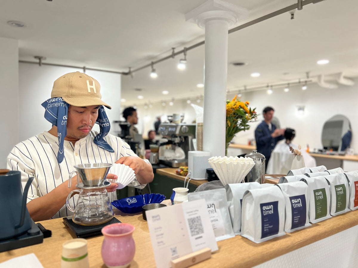 A barista wearing a beige cap with a blue Owntime bandana and a white pinstripe shirt prepares pour-over coffee at a counter lined with coffee bags and accessories in a bright barbershop