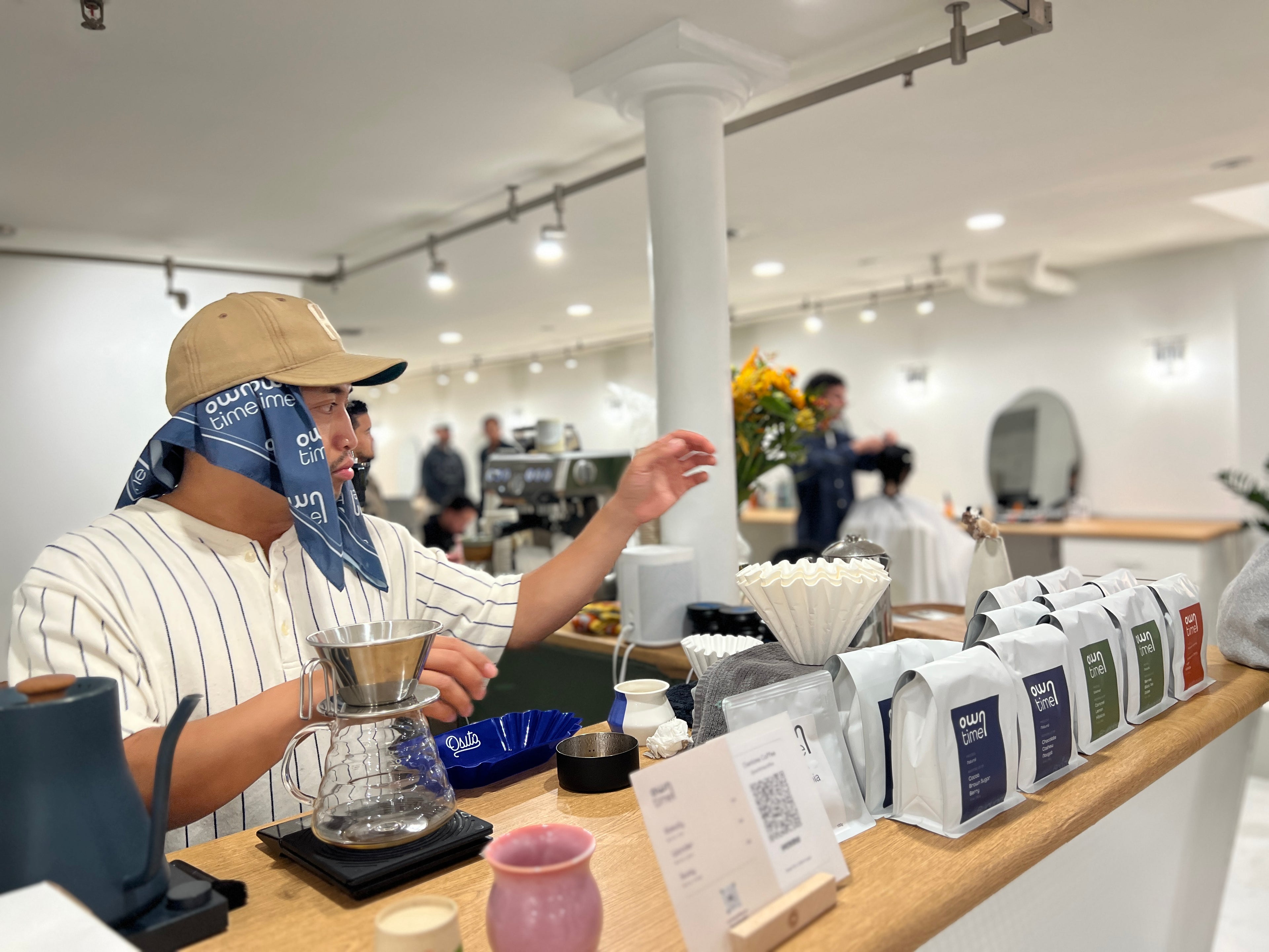 A person behind a coffee bar wearing a beige cap and blue Owntime Coffee bandana, pouring coffee using a pour-over setup on a wooden counter. Several packaged coffee bags are lined up in the foreground, with a bright, modern barbershop interior in the background.