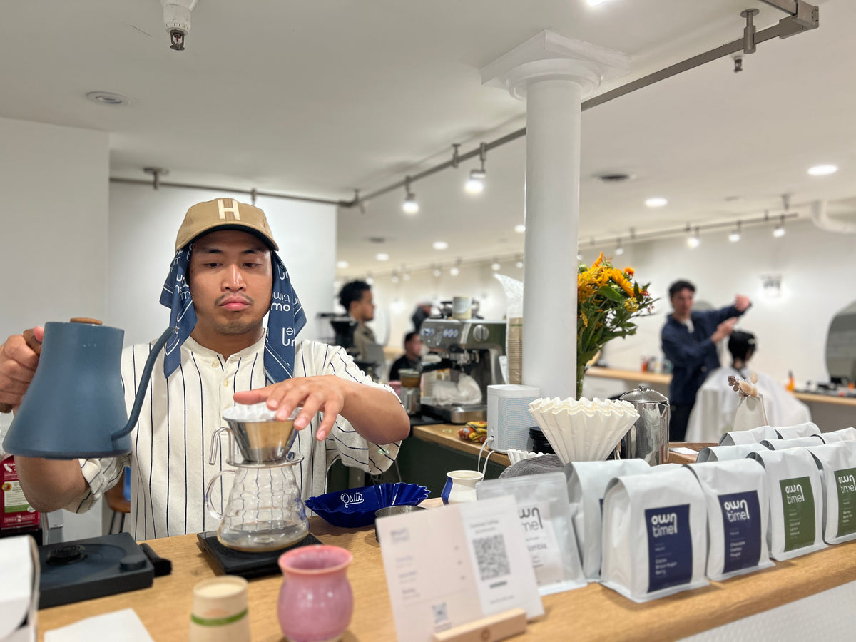 A barista wearing a beige cap and striped shirt pours hot water from a blue Fellow gooseneck kettle into a pour-over coffee dripper on a scale. In the foreground, several white bags of coffee with blue and green labels are lined up on a wooden counter in a bright, modern barbershop.