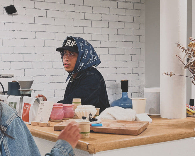 A person in a denim jacket and Owntime Coffee headscarf stands behind a wooden counter in a bright, modern barbershop with a white brick wall.