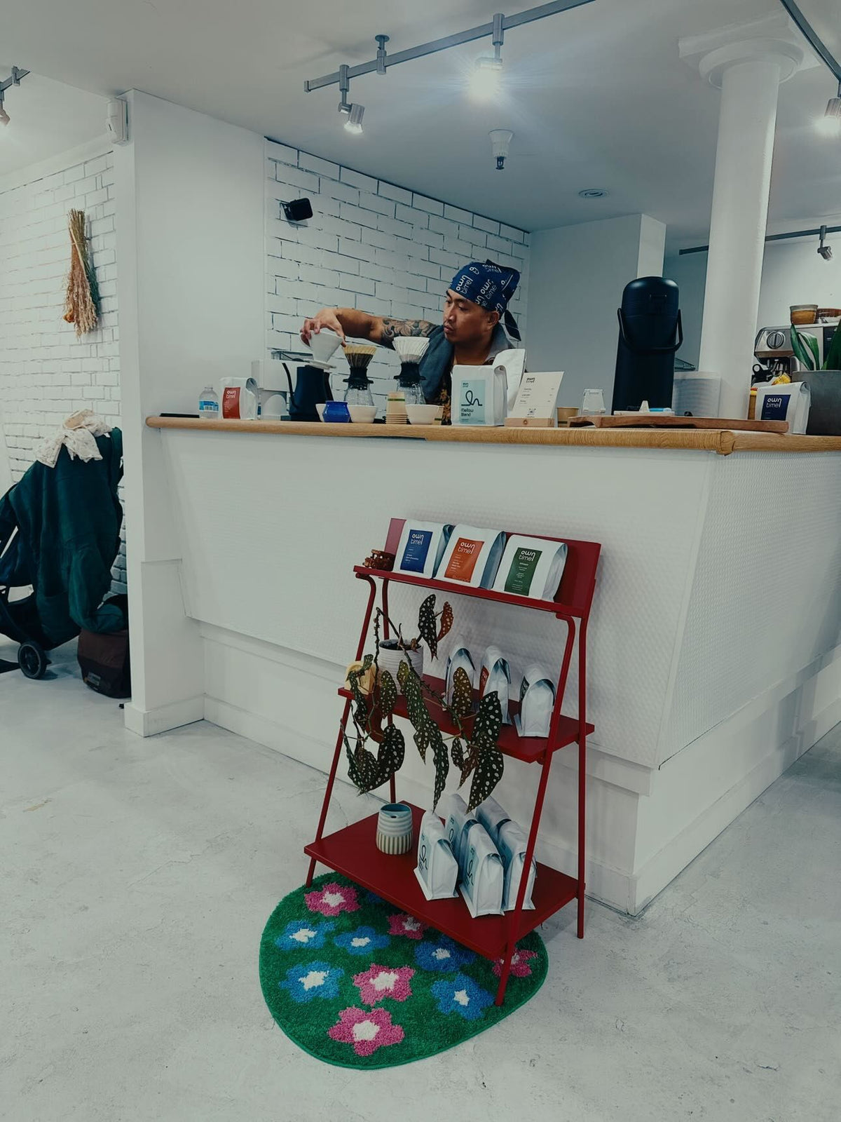 Person behind a coffee bar with a red display shelf featuring Owntime Coffee bags and accessories in a barbershop.