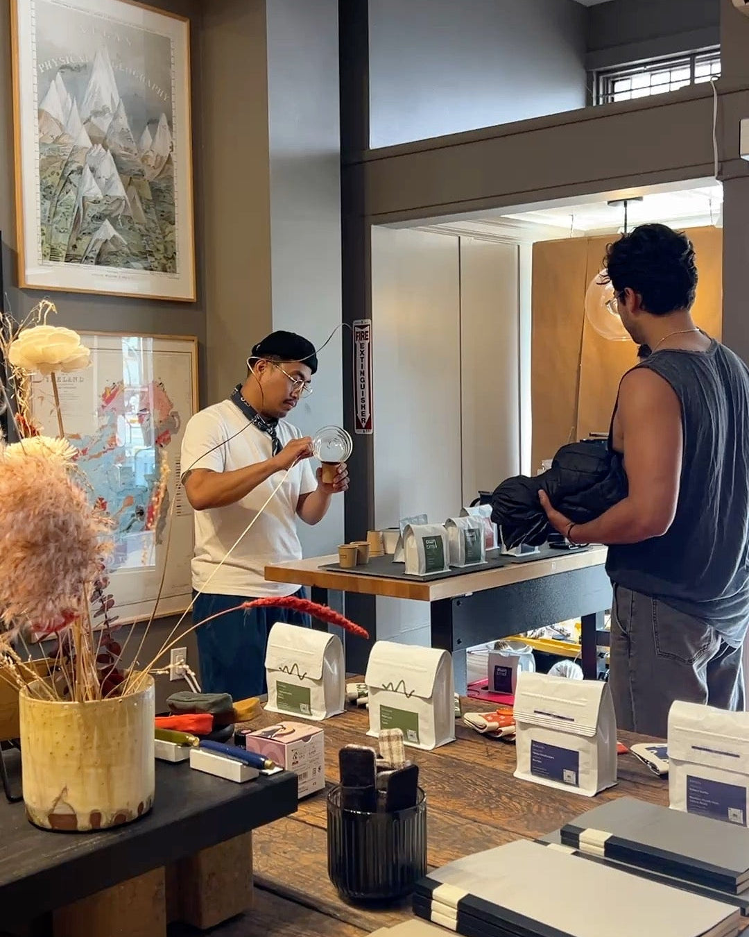 Owntime Coffee's roaster is pouring coffee from a carafe for a guest. In the foreground, a wooden table holds notebooks, sketchbooks, and a decorative dried flower arrangement in a beige pot.