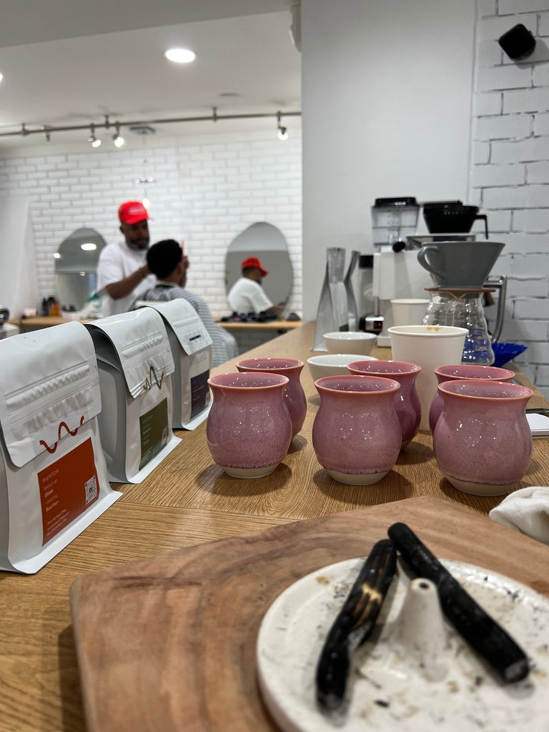 A wooden counter of a coffee bar displays several pink ceramic mugs, with white coffee bean bags lined up on the left and a blurred barbershop interior in the background.