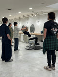 A lifestyle scene inside a modern, bright barbershop. Several people are gathered around a client sitting in a white salon chair, with one stylist demonstrating a haircut or styling technique.