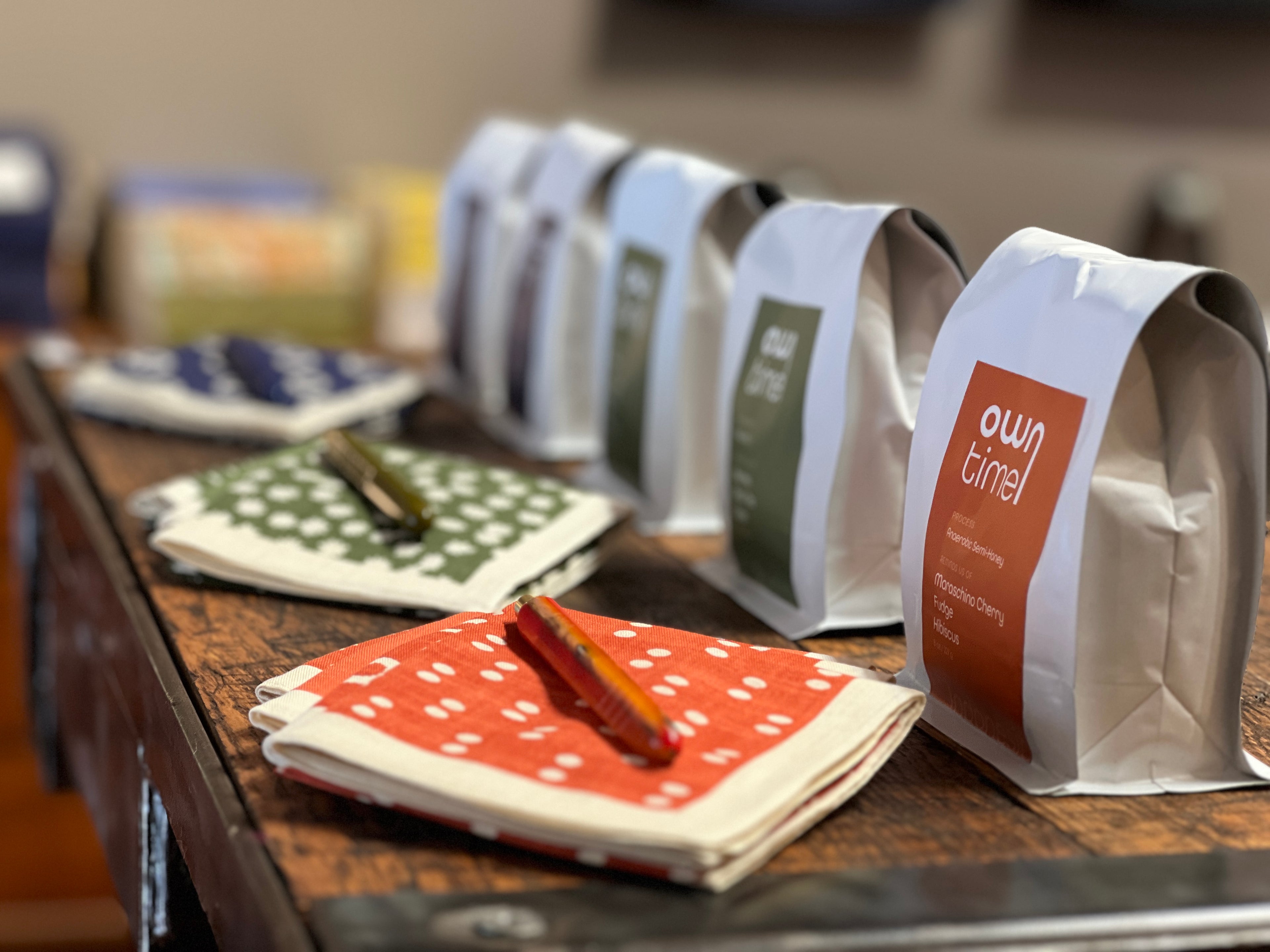 A row of Owntime Coffee bags resting on a rustic wooden table, styled with patterned handkerchiefs and pens from Topdrawer